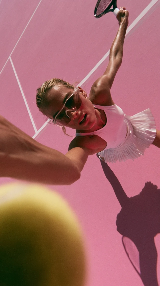 Dynamic cinematic editorial image of a female tennis player from a dramatic high angle perspective, reaching upward toward the camera on a vibrant pink tennis court with crisp white lines. Body extended in motion - one arm stretched toward the lens, the other holding a raised tennis racket - creating a powerful diagonal composition. A bright yellow tennis ball appears large and slightly out of focus in the foreground, emphasizing depth and movement. She wears a white fitted top and pleated skirt, sporty sunglasses, skin glowing under strong natural sunlight. Sharp shadows across the court add contrast and intensity. Background minimal and softly blurred. Wide lens exaggerates perspective; slight motion blur enhances realism. High contrast, warm tones, realistic skin texture, editorial sports campaign aesthetic, cinematic finish.