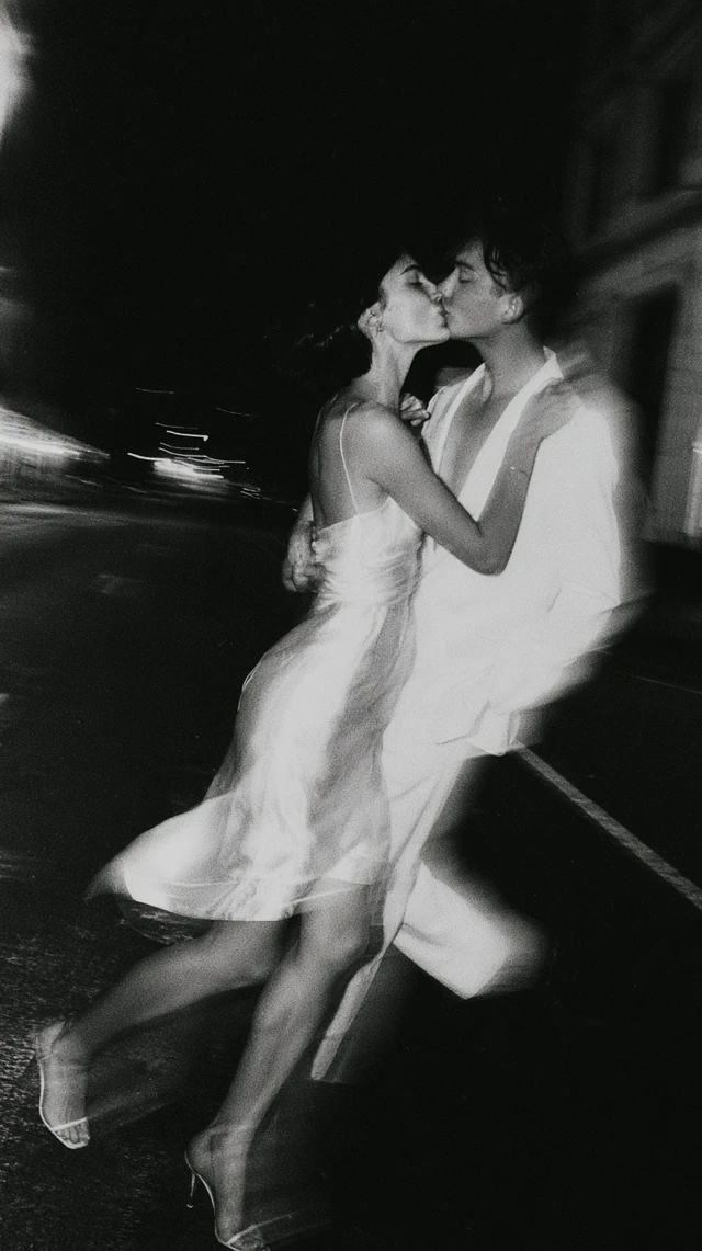 2026 modern romance in the city. A candid, high-fashion editorial black and white photograph of a very young gen-z couple in minimalist bridal attire sharing a breathless, blurry kiss while walking across an empty street, NY architecture in the background. Midnight, expansive pitch-black sky providing negative space at the top. Shot on Leica M4, 35mm Summilux f/1.4 lens, Kodak Tri-X 400 black and white film pushed to 3200 ISO. Vintage analog style, deep crushed and washed-out blacks, heavy cinematic film grain, subtle halftone newspaper texture, moody 1990s cult film aesthetic. Vogue Weddings vibe, raw, authentic, unposed, high contrast, harsh direct flash photography