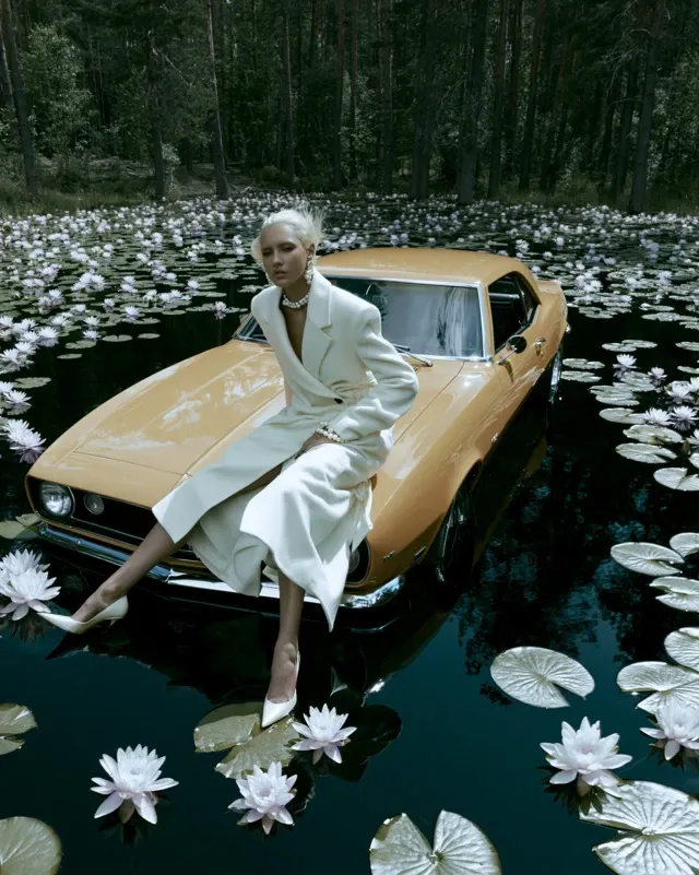 Tanned woman, platinum blonde hair in elegant updo or windswept look, perching confidently on a vintage retro yellow orange Chevrolet Camaro half submerged in a dark mysterious forest lake - roof and hood barely above water with shimmering reflections. She wears a structured cream/ivory wool coat dress, pearl accessories, pointed heels. Dramatic pose: one leg extended, body twisted, intense commanding gaze into the distance. Lake covered in blooming water lilies and lotus flowers, large lily pads on dark teal water. Tall ancient pine forest surrounds lake, moody misty light filtering through trees. Slightly low angle, full figure visible, cinematic surreal editorial fashion photography. Rich deep greens and dark teals contrast warm orange car and pale outfit. Cool neutral color balance. No orange cast, amber tones, golden hour lighting, teal orange grading, glossy commercial beauty, or plastic skin. Realistic skin texture, visible pores, tactile surface detail, restrained palette, unconventional composition, editorial print feel.
