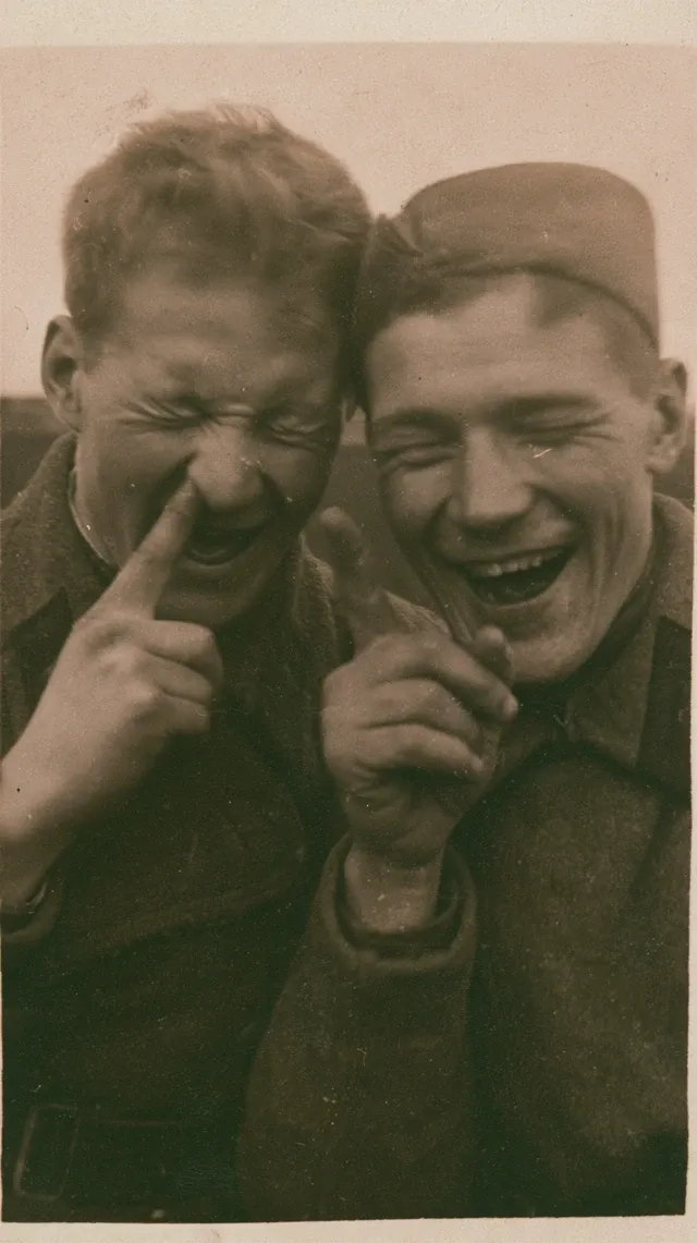 Realistic photograph of two Russian soldiers laughing. They are standing with their eyes closed, pointing at someone off-frame, and laughing. The day is cloudy and humid. The soldiers are seen from the waist up. The photograph is current, taking place in the present.