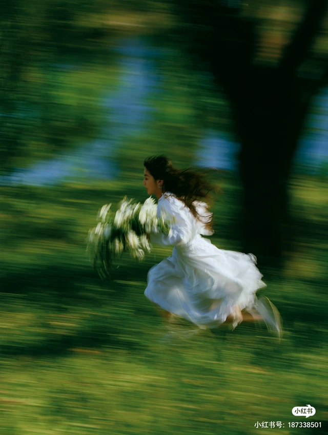 a person in a white dress runs through a field of green grass, holding white flowers. the shot is motion blurred, conveying movement. blurred green and blue elements, likely foliage and water, form the background, along with a dark tree trunk. chinese text is visible in the bottom right corner: "小红书" and "小红书号: 187338501".