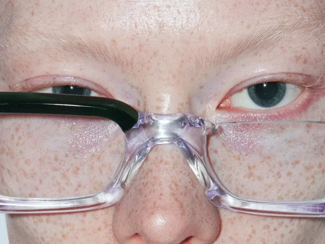 [IMAGE URL] extreme macro close-up portrait of an adult person’s eyes and nose bridge, direct frontal gaze, dark blue-green eyes, rectangular eyeglasses with a glossy black upper frame and transparent lower rims, densely freckled fair skin, visible pores and fine facial hairs, subtle moisture beneath the eyes, perfectly centered symmetrical composition, tightly cropped face filling the entire frame, harsh direct on-camera flash, crisp specular reflections on the lenses, raw editorial beauty photography, intimate and slightly unsettling mood, photorealistic, ultra-detailed natural skin texture, sharp focus, high contrast, warm neutral color grading, no retouching, no background visible