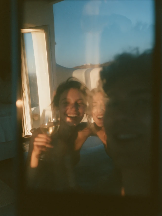 close-up of a phone screen in a dark bedroom showing a glamorous couple laughing and sipping wine in a bathtub on a sun-drenched terrace overlooking the sea in Mykonos, white architecture, golden light on screen contrasting with dark room, young man's tired face faintly reflected in the glass, late night, cinematic, 35mm film look, cold blue tones ar 16:9 style raw