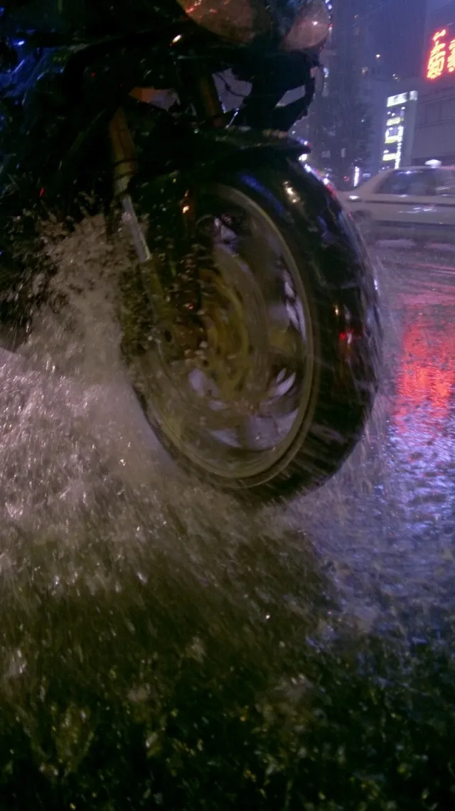 Extreme low-angle close-up of the front wheel of a black sport motorcycle racing through rainy Tokyo at night. The wheel dominates the frame. Water sprays violently from the tire across the wet asphalt. Red neon signs reflect on the spinning rim and rain-soaked road. Cars blur past in the background. Strong motion blur, intense speed, cinematic neo-noir atmosphere, ultra realistic, action movie frame.