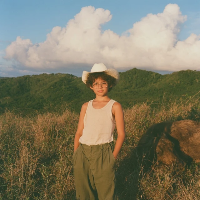 medium shot of a young boy with light brown skin, curly brown hair, and wire rimmed glasses, wearing a white cowboy hat, a sleeveless white tank top, and olive green trousers. he stands in a grassy field, hands in his pockets, looking toward the camera. in the background, rolling green hills under a blue sky filled with puffy white clouds. a brown water buffalo lies on the ground to his right in the tall dry grass. the scene is illuminated by warm, golden hour sunlight.